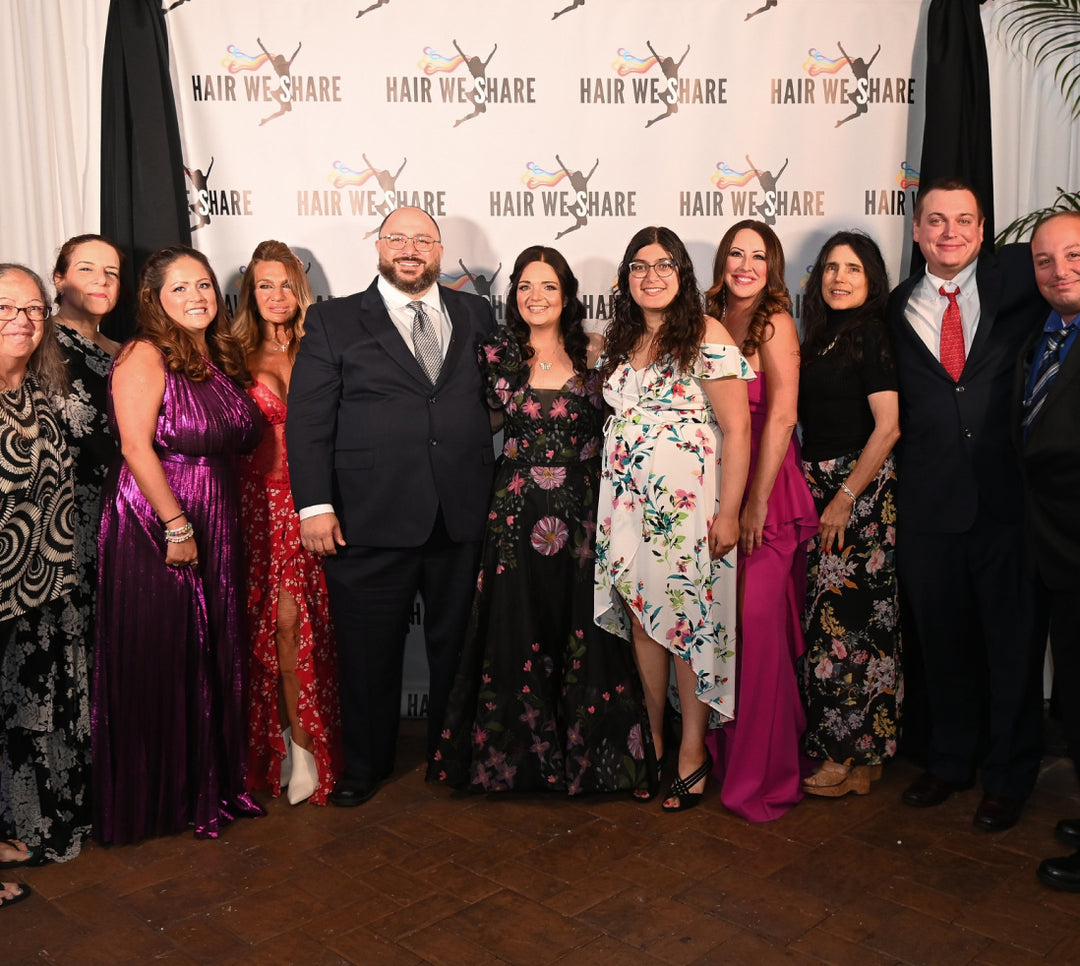 Group of people in dress attire posing for a photo against a Hair We Share backdrop at the 12th annual Hair We Share gala.
