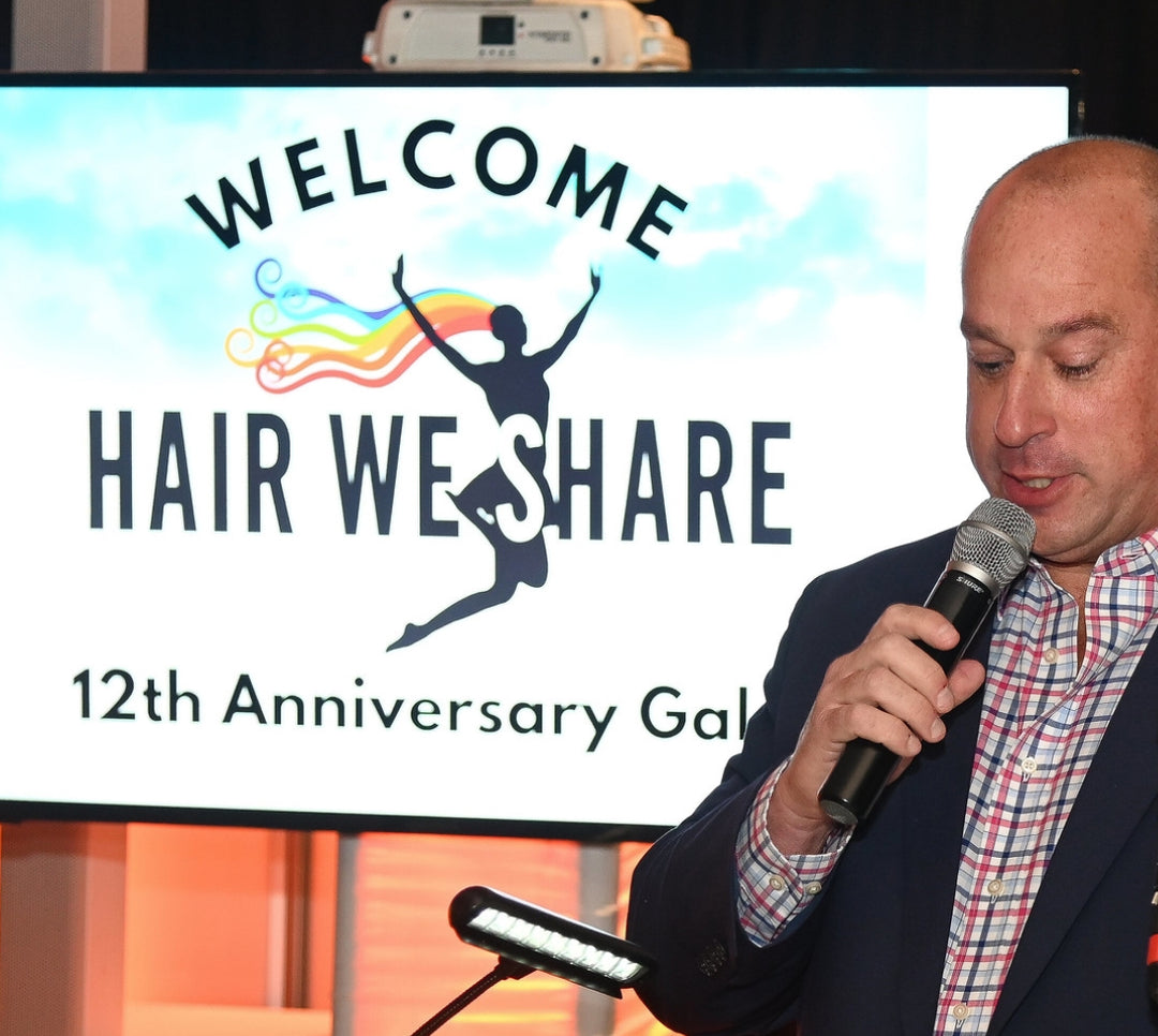 Man speaking into a microphone with 'Hair We Share' 12th Anniversary Gala sign in the background